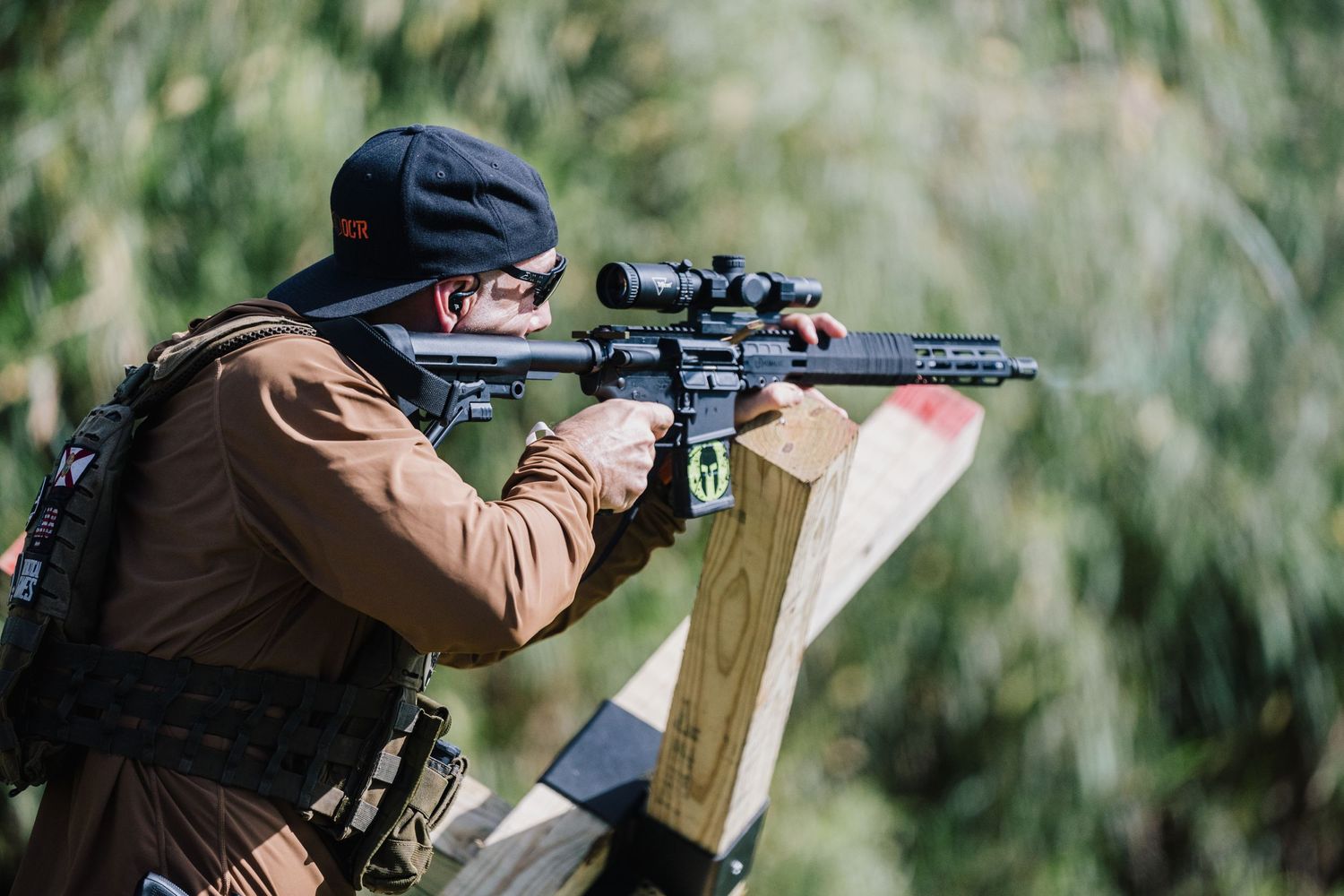 Shooter in tactical gear aims rifle at outdoor shooting range.