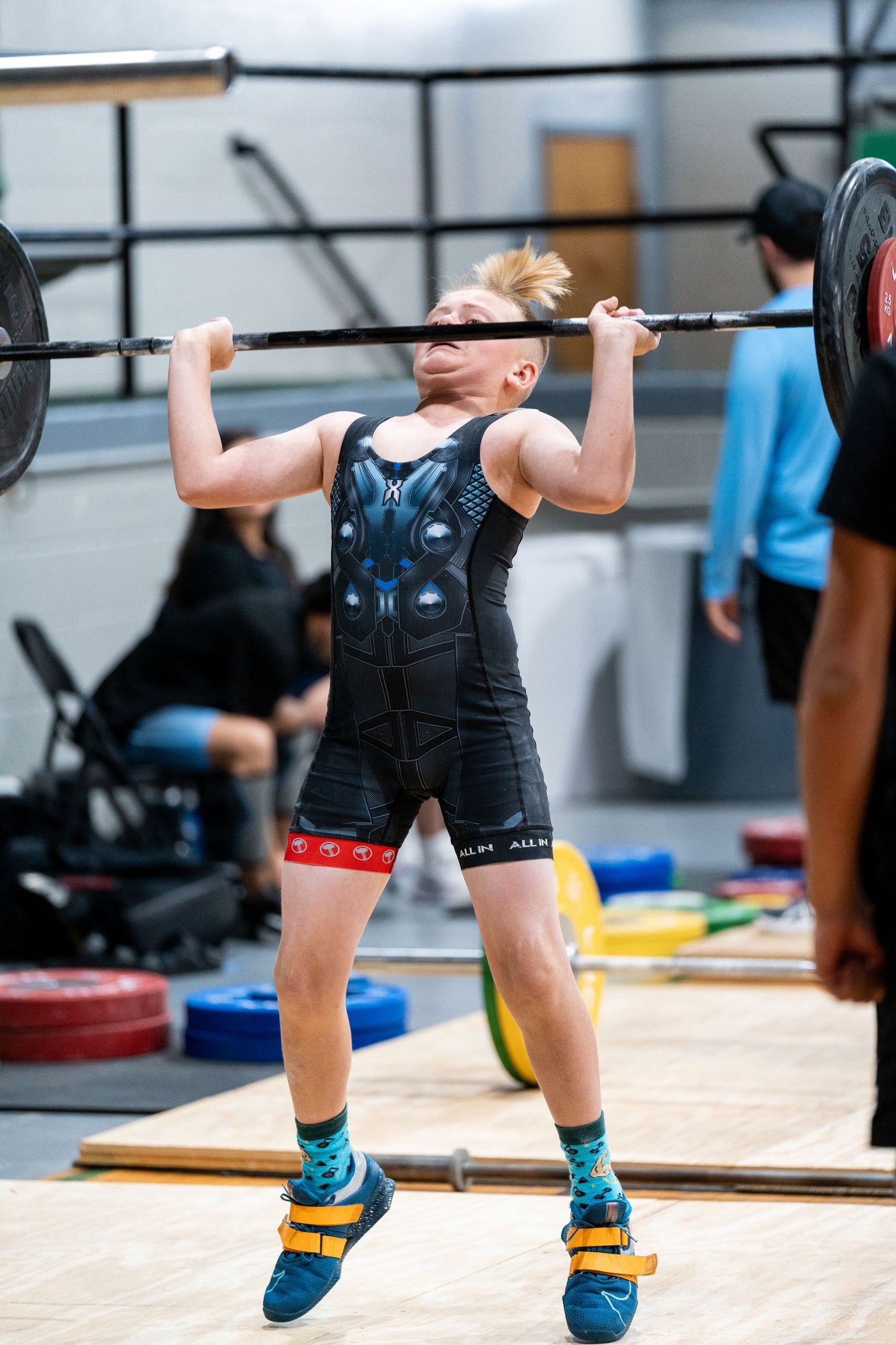 Young athlete executes an overhead lift with barbell while wearing blue shoes and black singlet.