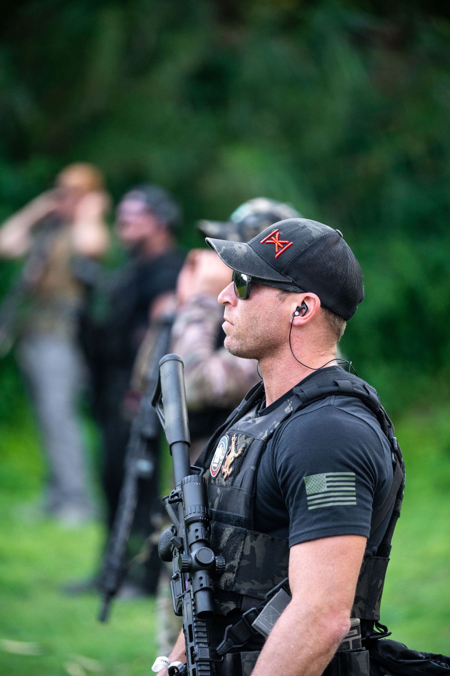 Tactical instructor in black gear and baseball cap stands ready at outdoor range.