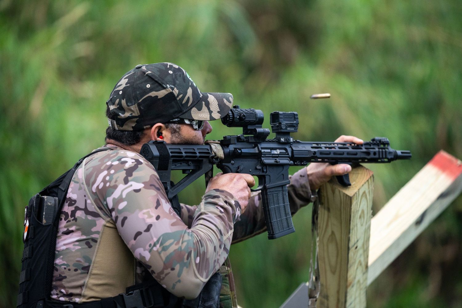 Shooter in camouflage gear aims rifle from wooden shooting position.