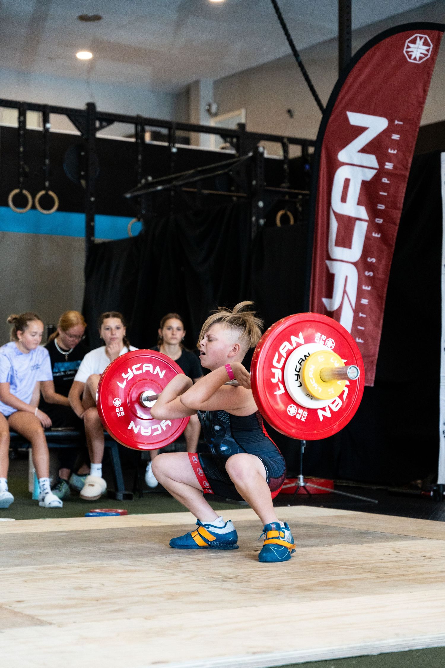 Young athlete performs an Olympic weightlifting squat with red plates on wooden platform next to a red banner.