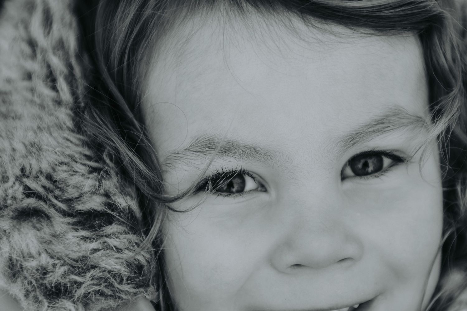 Close up black and white portrait of a smiling child with bright eyes and dark hair against a fluffy textured background.