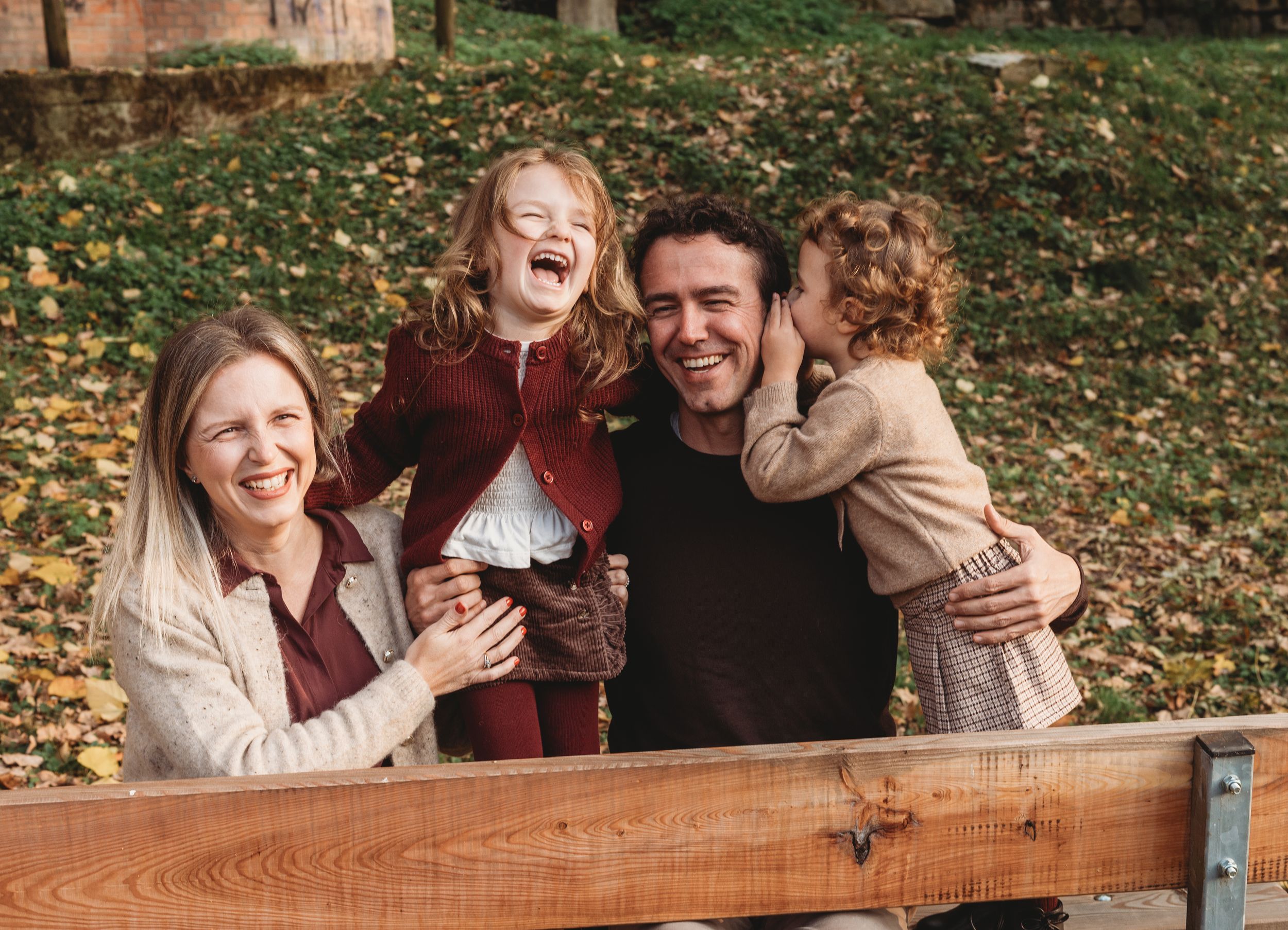 A joyful group laughs together while standing behind a wooden fence on an autumn day with fallen leaves in the background.