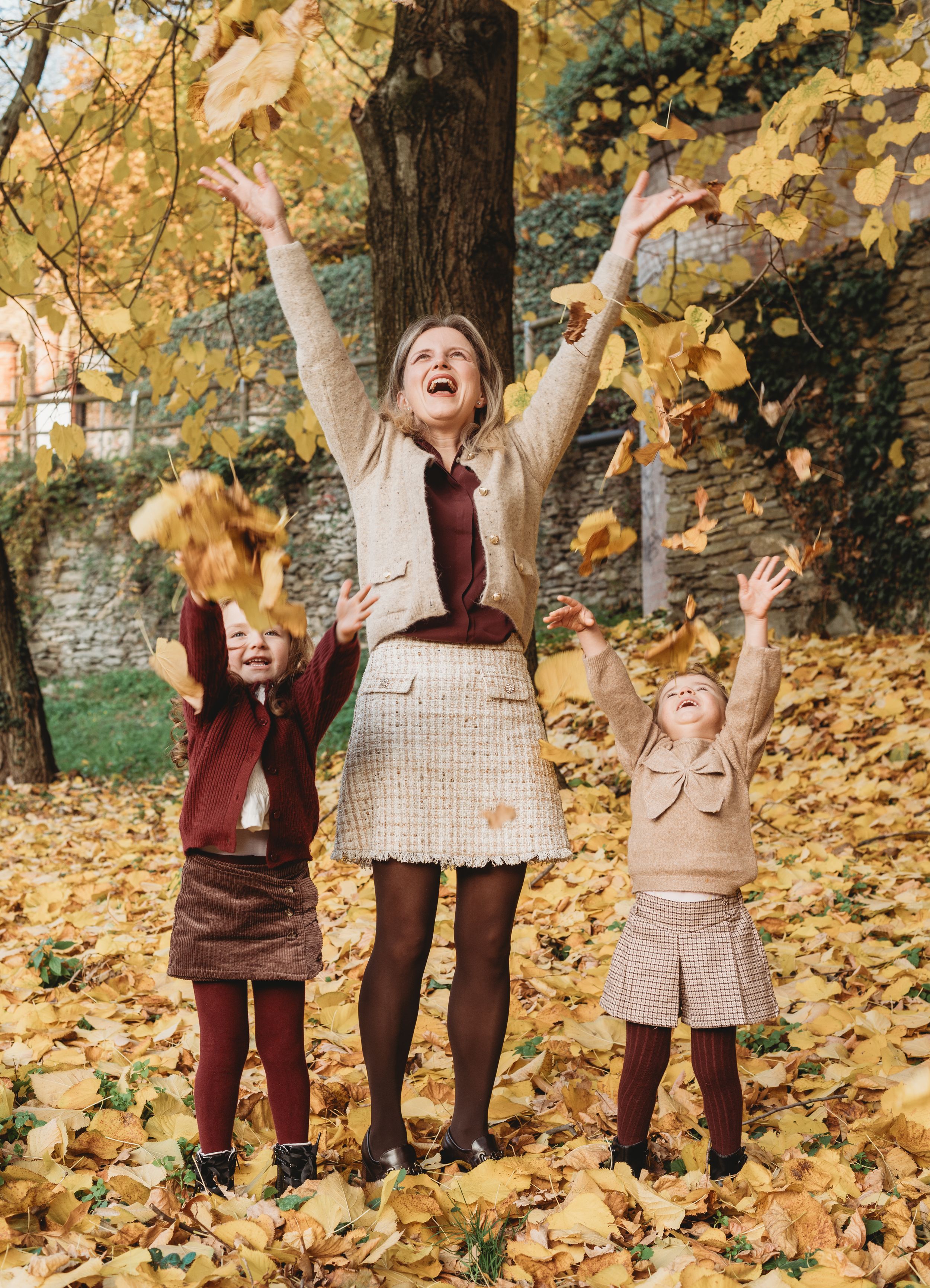 Three people in autumn outfits joyfully toss fallen leaves in the air while standing on a carpet of yellow foliage.