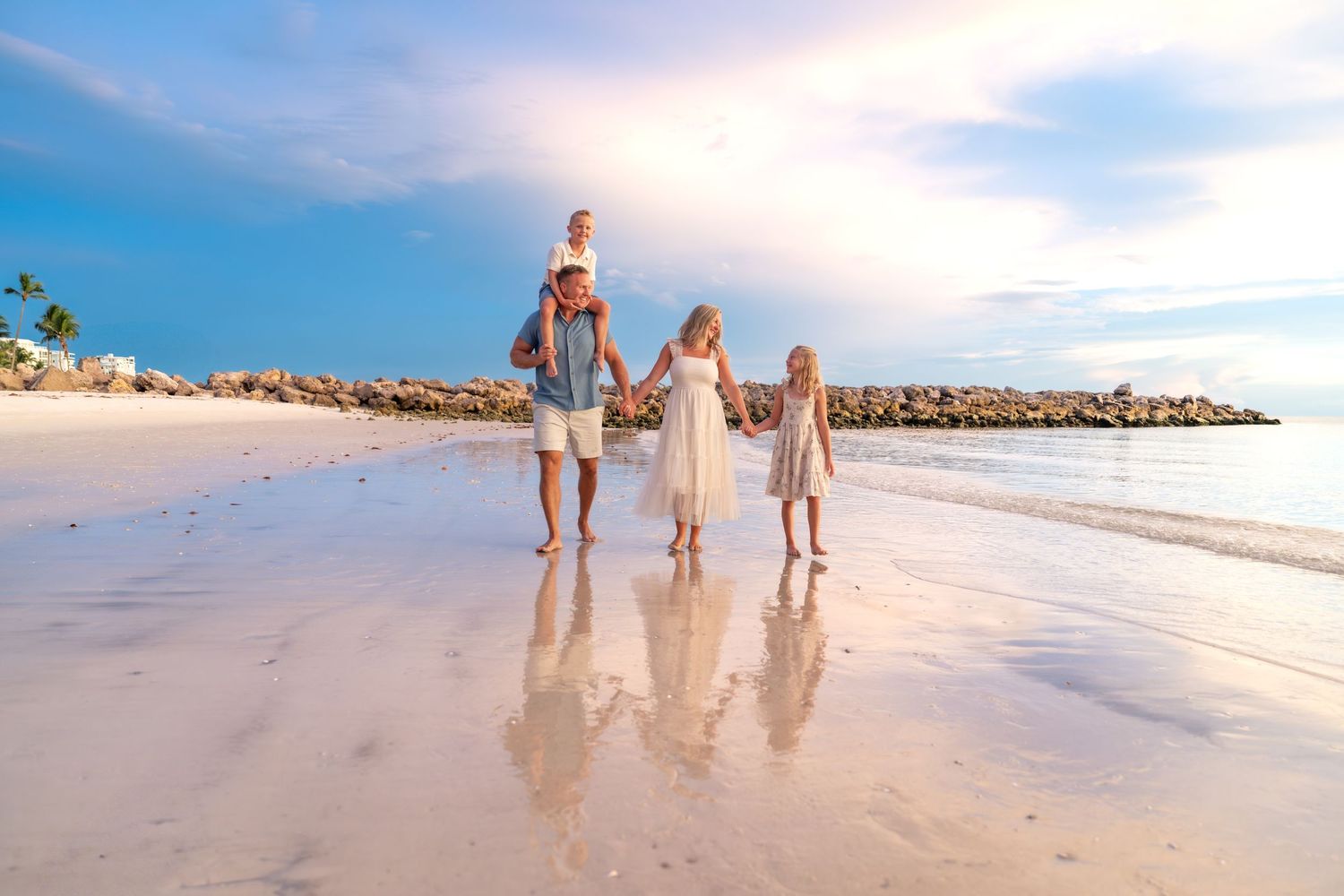 Four people in white outfits sitting on a beach at sunset with ocean waves in background.