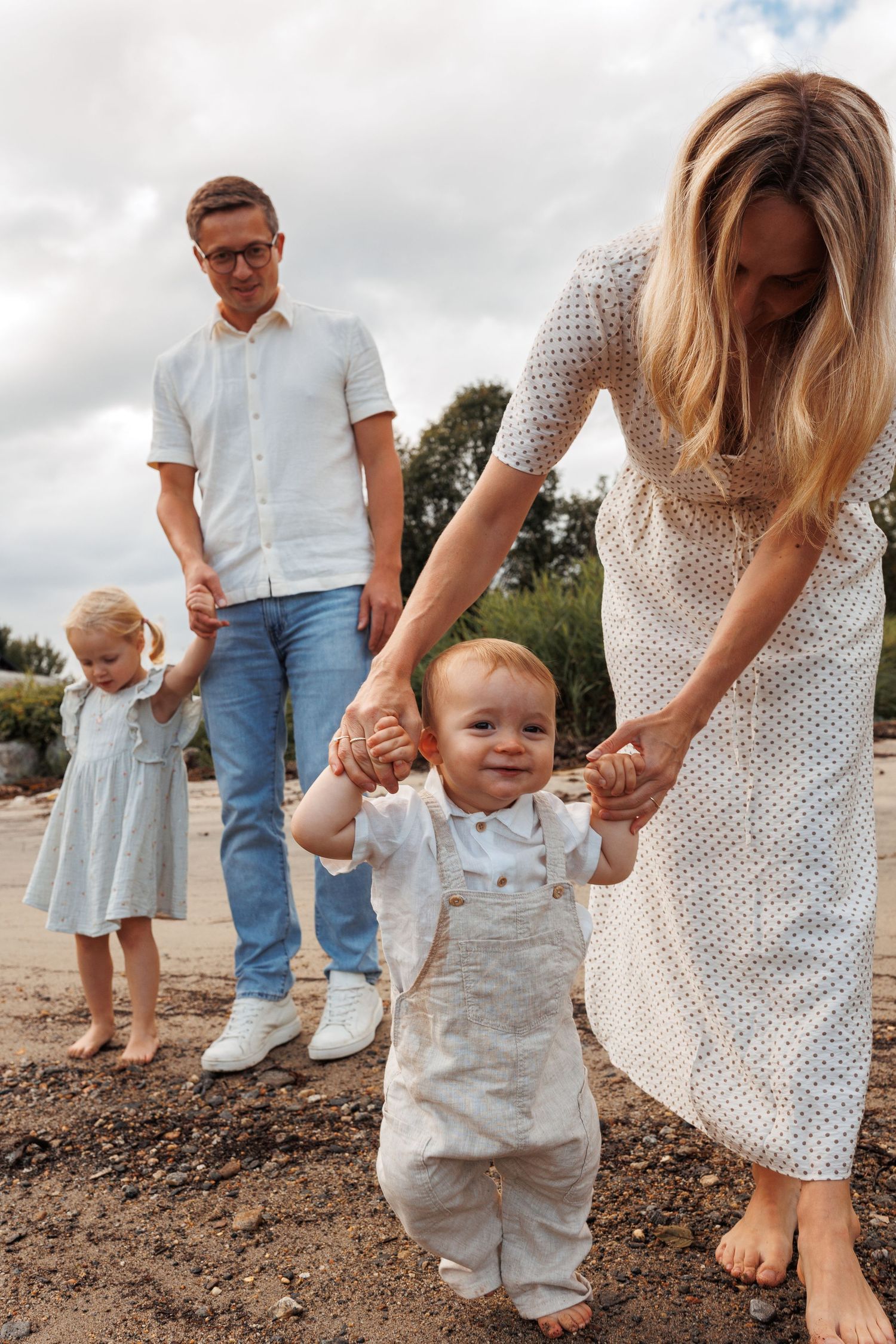 A family stands together on a dirt path as a small child takes joyful steps while being held by their parents.