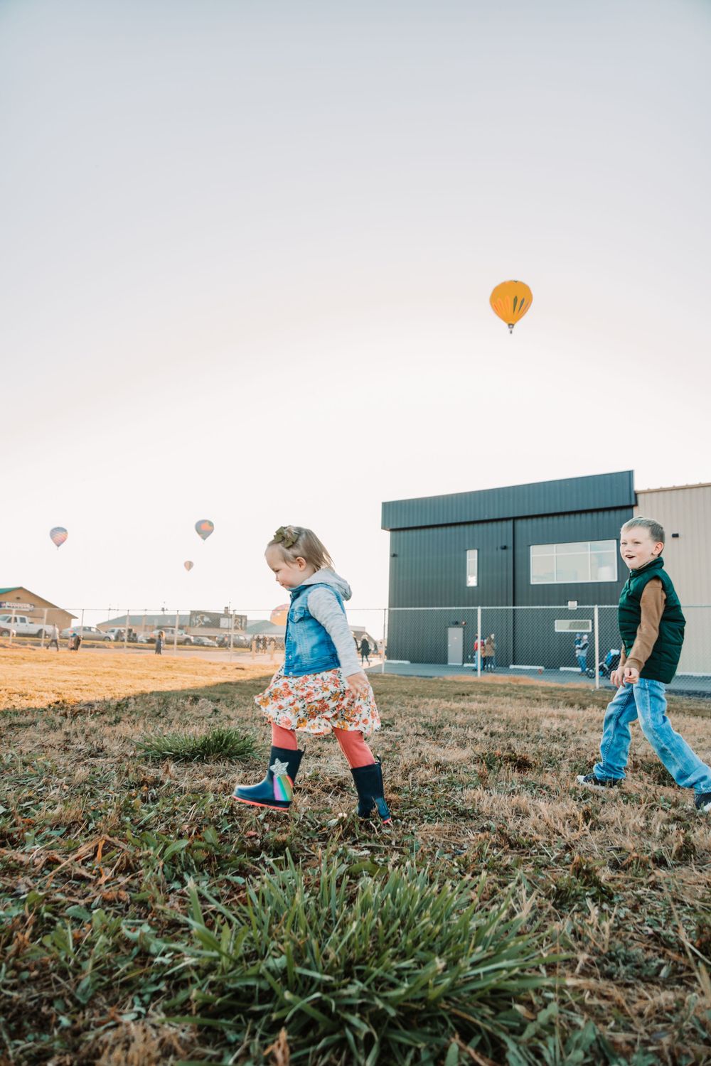 Children walking on grassy hill with hot air balloons floating in distant sky.
