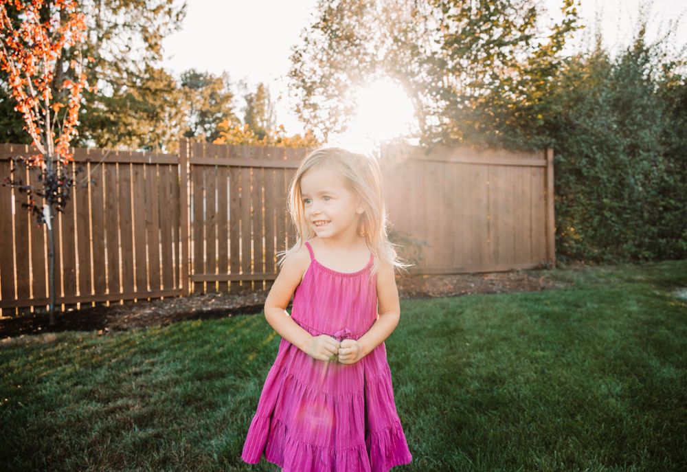 Child twirls in pink dress as sun sets behind wooden fence in backyard.