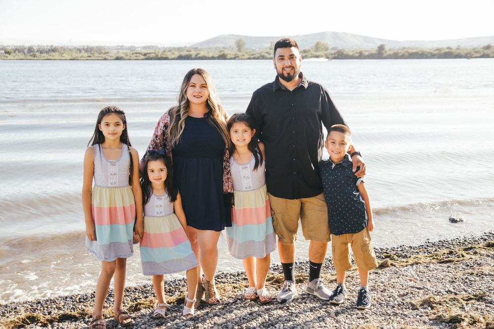 Family group stands together on rocky beach shore with water and mountains in background.