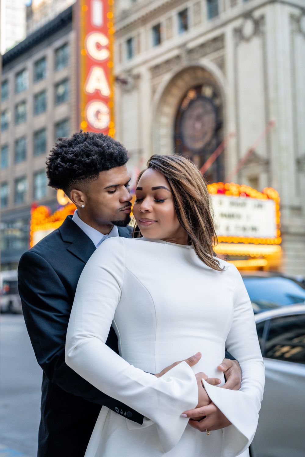 Romantic couple embraces in front of illuminated city buildings at night.