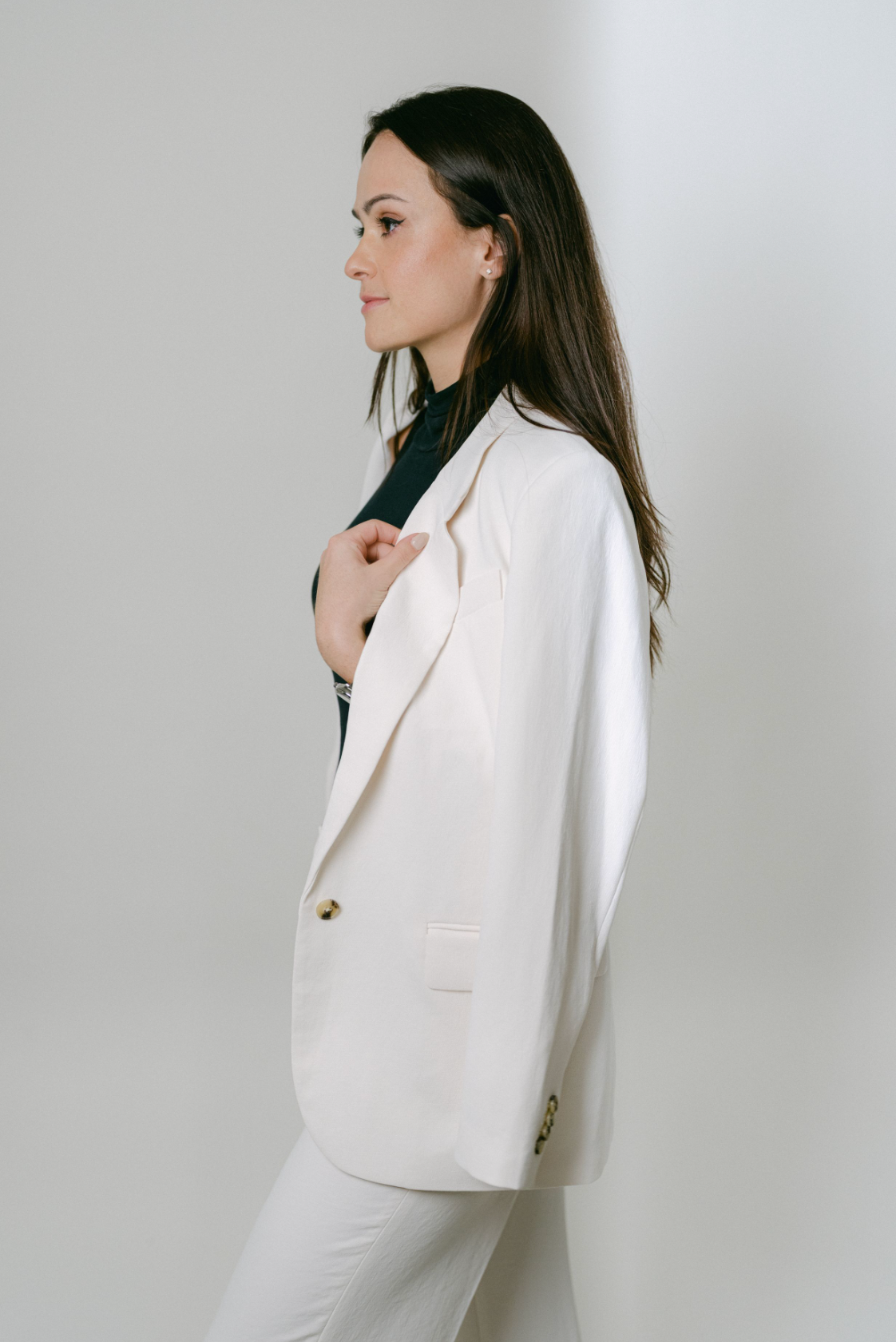 A side profile studio portrait featuring a model in an elegant white blazer and dark top against a light background.