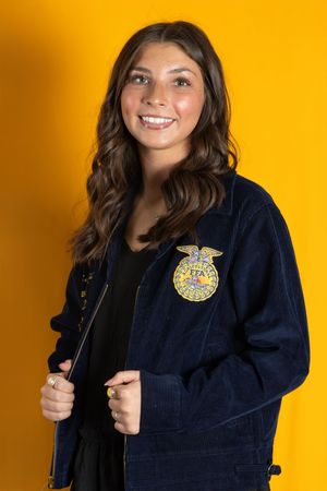 Young woman smiling in navy FFA jacket with emblem on yellow background. Salem and Gloucester County Senior Portraits