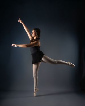A ballet dancer in black leotard performs an arabesque on pointe shoes against a dark studio background. Salem and Gloucester County Senior
