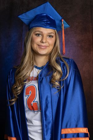 Young woman in blue graduation cap and gown smiling, wearing a sports jersey underneath for her senior portrait.