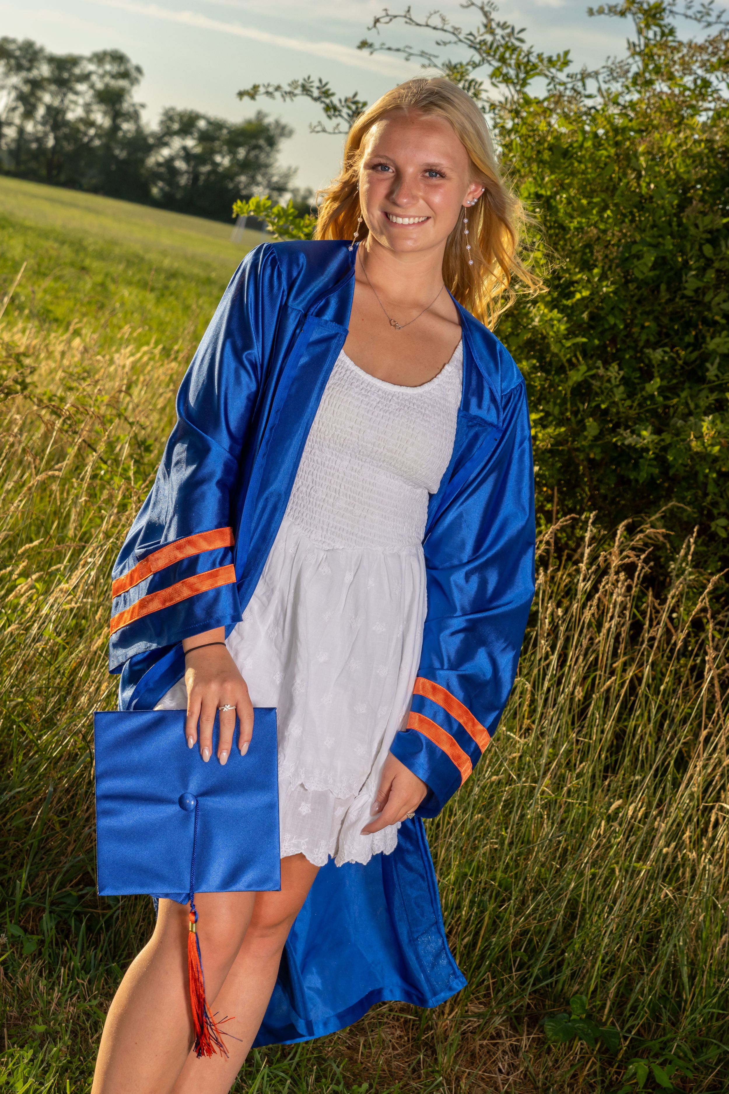 Smiling blonde woman in blue graduation gown with orange stripes poses outdoors in a sunny green field.