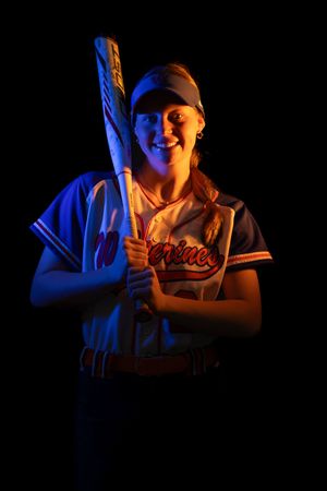 A senior softball player Woodstown poses with a bat against a dark background with dramatic blue and orange lighting.