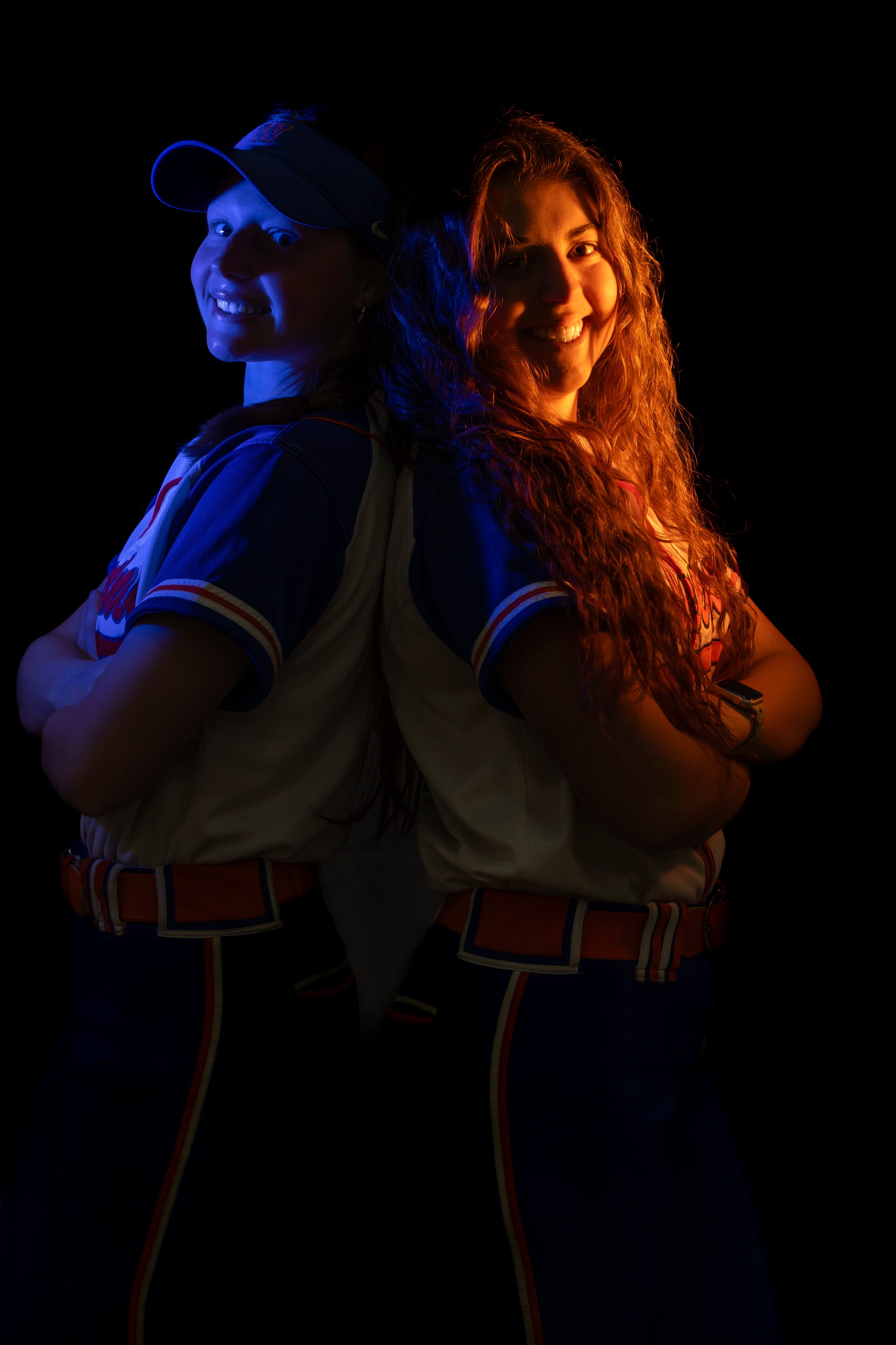 2 softball players from Woodstown High school stand back-to-back in dramatic lighting with blue and orange tones against a dark background.