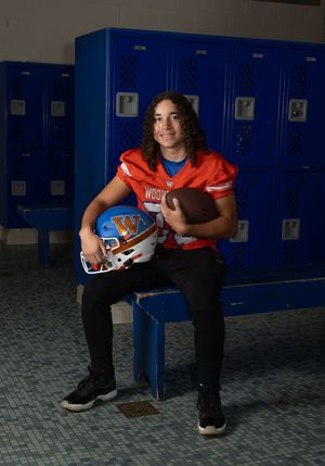 High school football player in orange jersey sits in locker room holding helmet and football. Salem and Gloucester County Senior Portraits