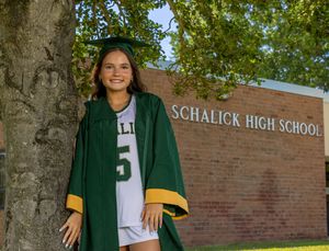 High school graduate in green cap and gown poses by a tree in front of Schalick High School brick building. SJ Senior Portaits