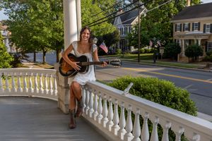 Woman in white dress playing acoustic guitar on a charming porch with American flags and tree-lined street. South Jersey Senior Portraits
