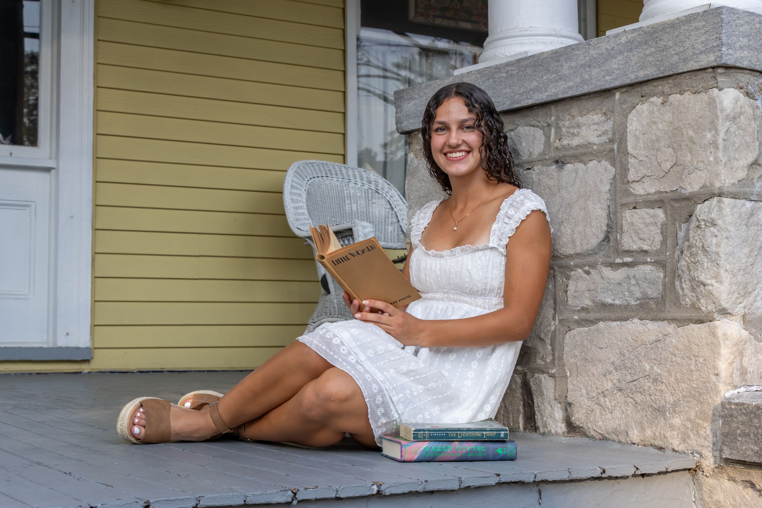 Young woman in white dress sitting on porch steps, smiling and holding a book with stacked books beside her.  south jersey Senior Portraits