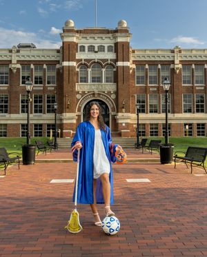 A female graduate in blue cap and gown poses with a lacrosse stick and soccer ball in front of a brick university building. Senior Portraits