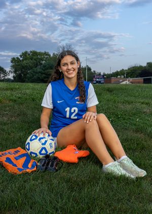 A smiling girl in a blue soccer jersey number 12 sits on grass with a soccer ball and cleats. Salem and Gloucester County Senior Portraits