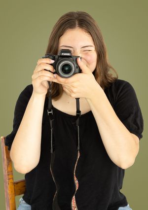 Young woman in black top smiling while taking a photo with a camera against olive background. Salem and Gloucester County Senior Portraits