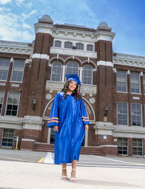 Graduate in blue cap and gown poses proudly in front of a brick school building on a sunny day. Salem and Gloucester County Senior Portraits