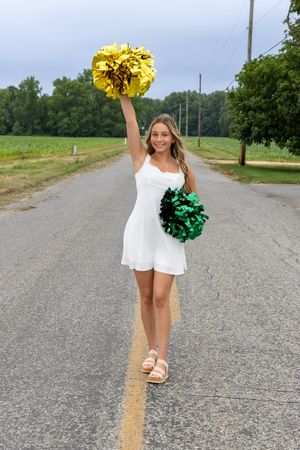 Cheerleader in white dress holding gold and green pom-poms on a rural road with green fields and trees. SJ Senior Portraits