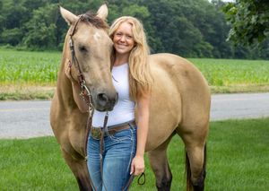 Young blonde woman smiling beside a buckskin horse on a green farm with cornfields in the background. Salem County Senior Portratits