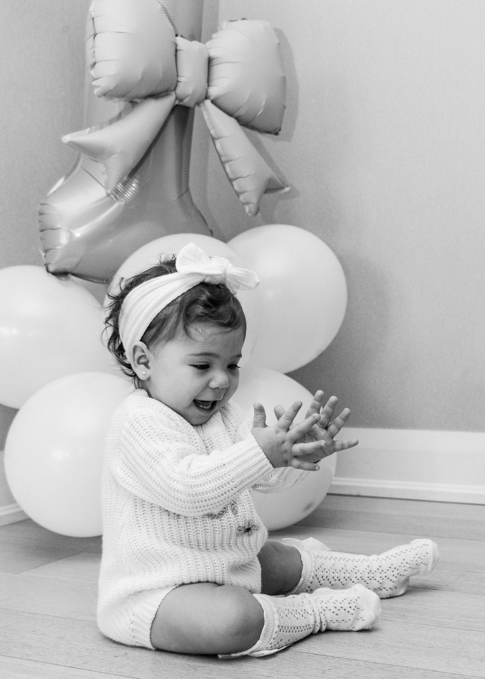 A baby in a white knit dress sits on a wooden floor surrounded by light gray balloons and bows.