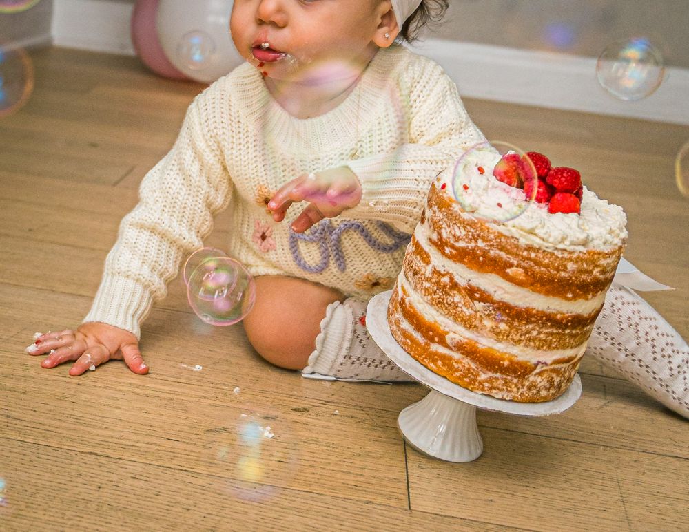A small child in a white sweater reaches for a homemade birthday cake with strawberries on top.