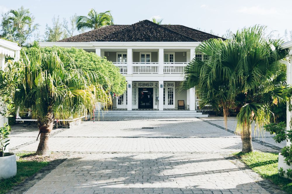 Elegant white colonial-style mansion with palm trees lining a paved driveway, surrounded by lush tropical greenery.