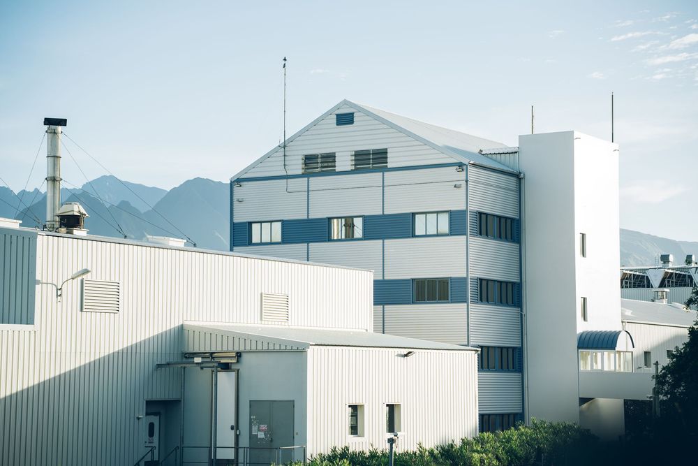Large white industrial factory building with multi-story facade, metal structures, and mountain backdrop under clear sky.