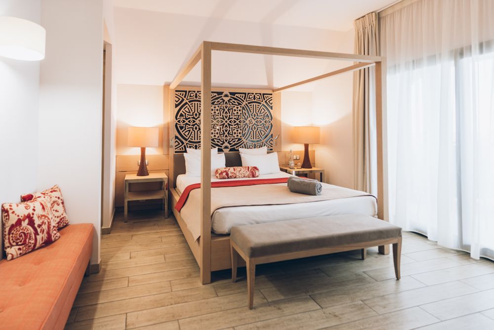 Modern hotel room interior with wooden floors, white bedding, and natural light streaming through sheer curtains.