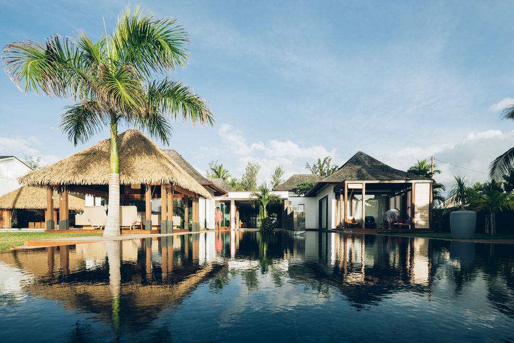 Tropical resort pool reflecting thatched-roof bungalows and palm trees under a blue sky.