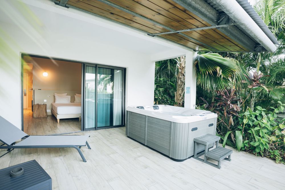 Luxurious outdoor patio area with hot tub, lounge chair and tropical plants adjacent to modern bedroom suite.