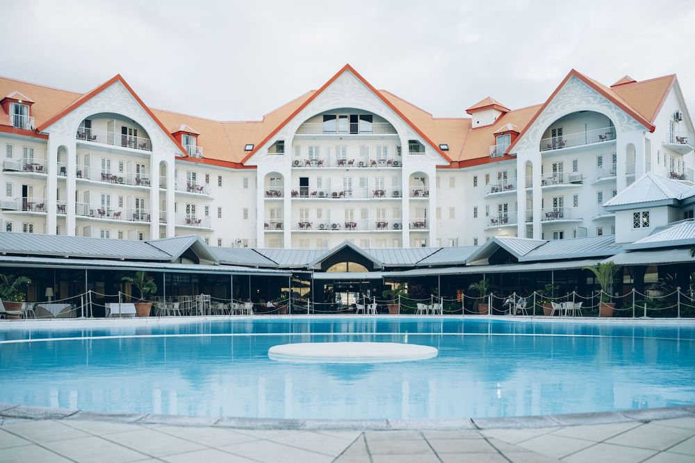 Elegant white resort hotel building with blue swimming pool and beach-style architecture reflecting in calm waters at dusk.