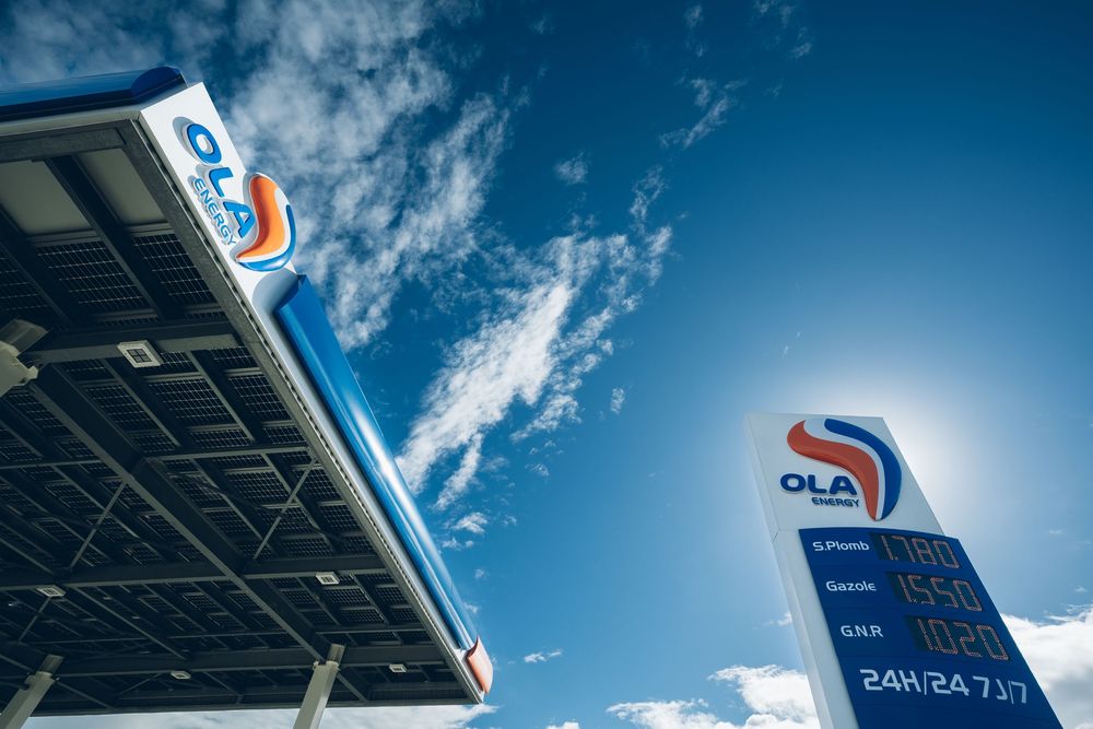 OLA fuel station with price sign displaying petrol and diesel prices under a bright blue sky with solar panels.