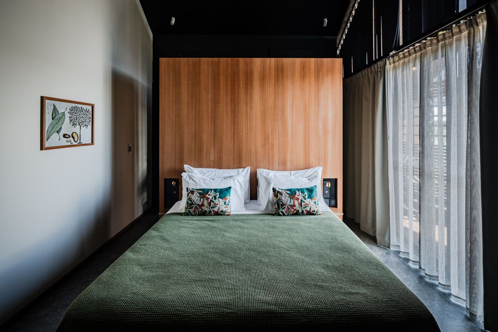 Modern hotel bedroom with green bedspread, white pillows, wood panel headboard, and sheer curtains.