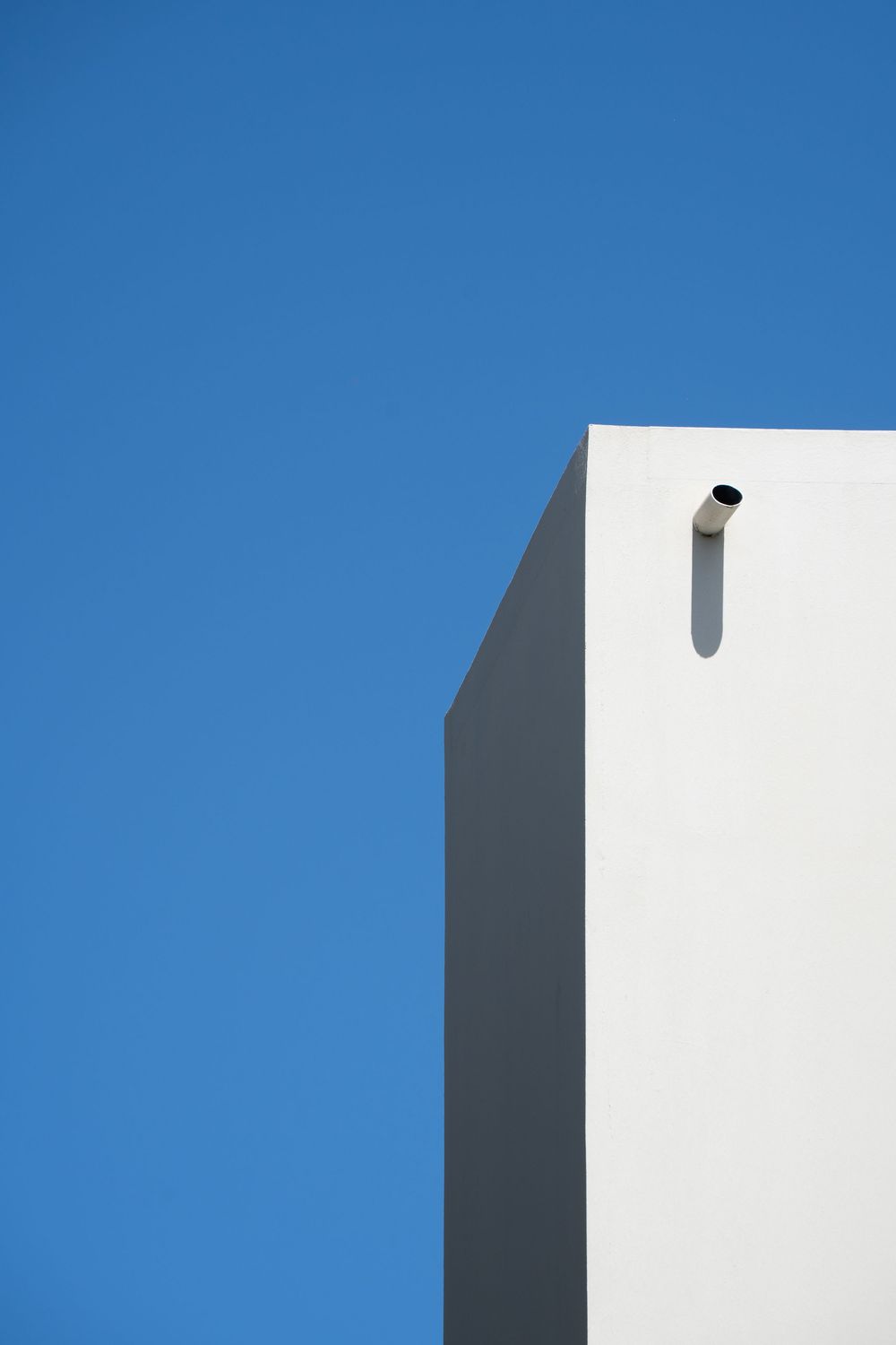 Minimalist white building corner with a small pipe against a clear blue sky.