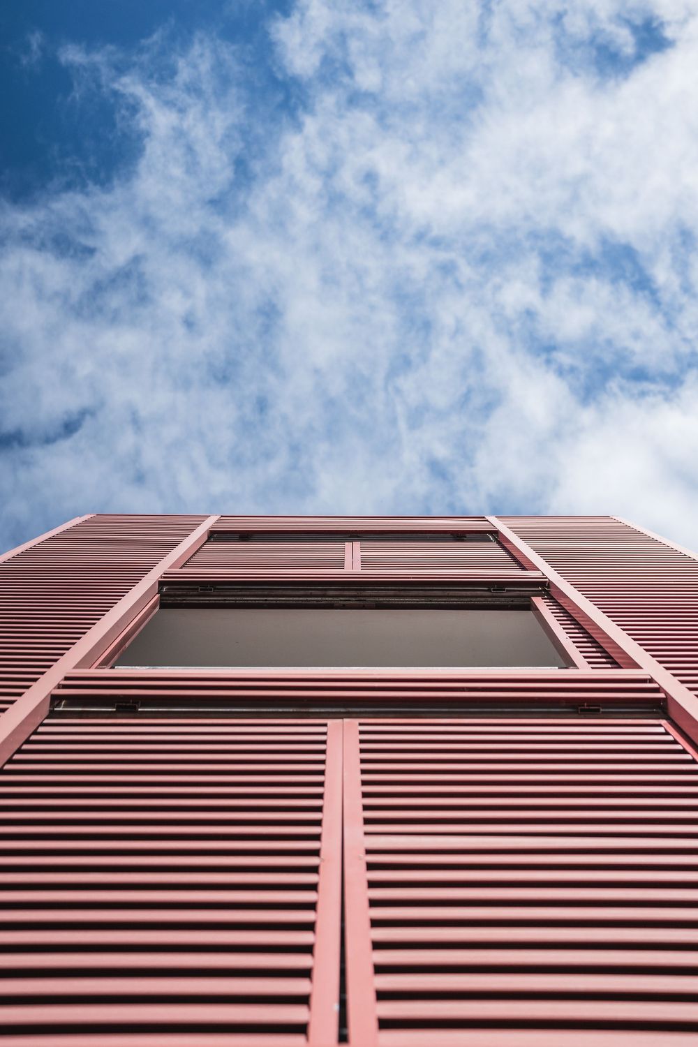 Low angle view of a modern red building facade with horizontal louvers and a window against a blue cloudy sky.