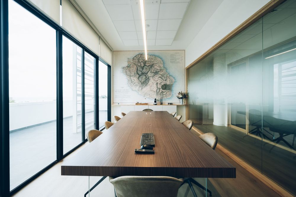 Modern corporate boardroom with long wooden table, white chairs, glass walls, and a world map artwork in natural light.