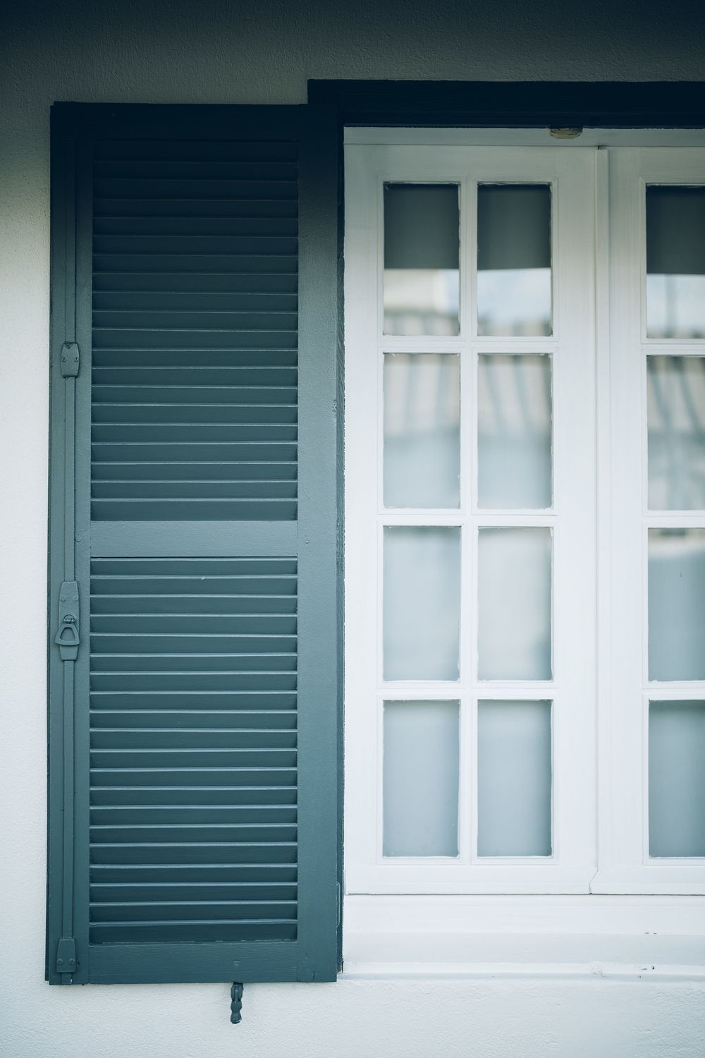 Dark green louvered window shutter beside white French window on a white wall exterior.