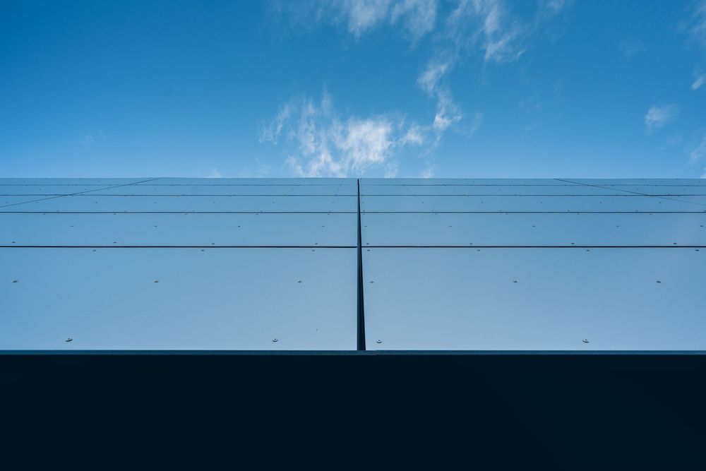 Minimalist blue sky with wispy clouds above a dark wall with horizontal lines and a vertical pole.