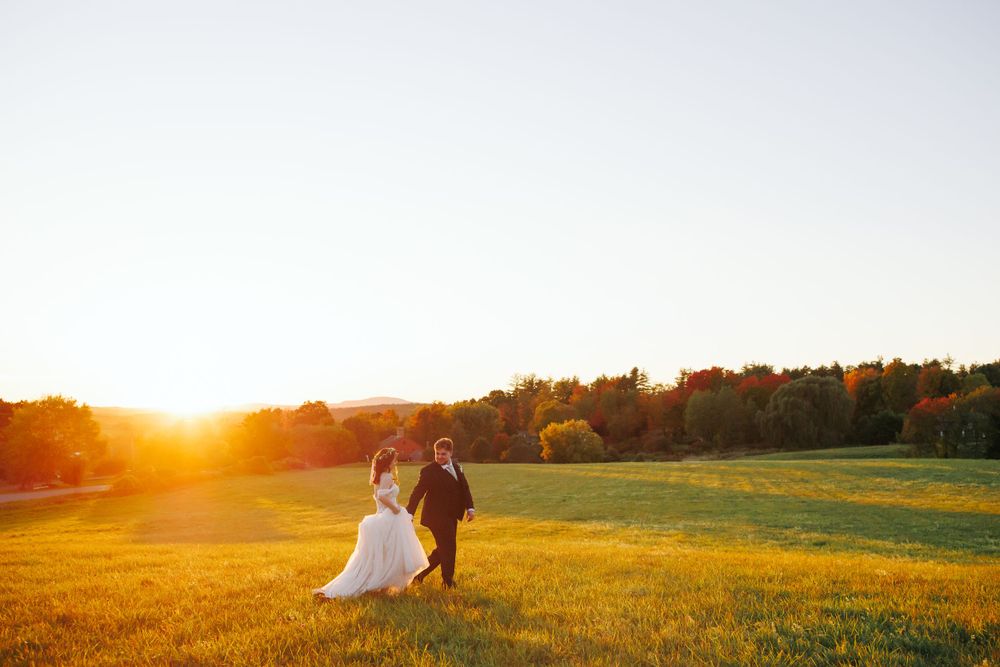 A couple walks hand in hand across a golden farmstead field at sunset in Harvard, MA