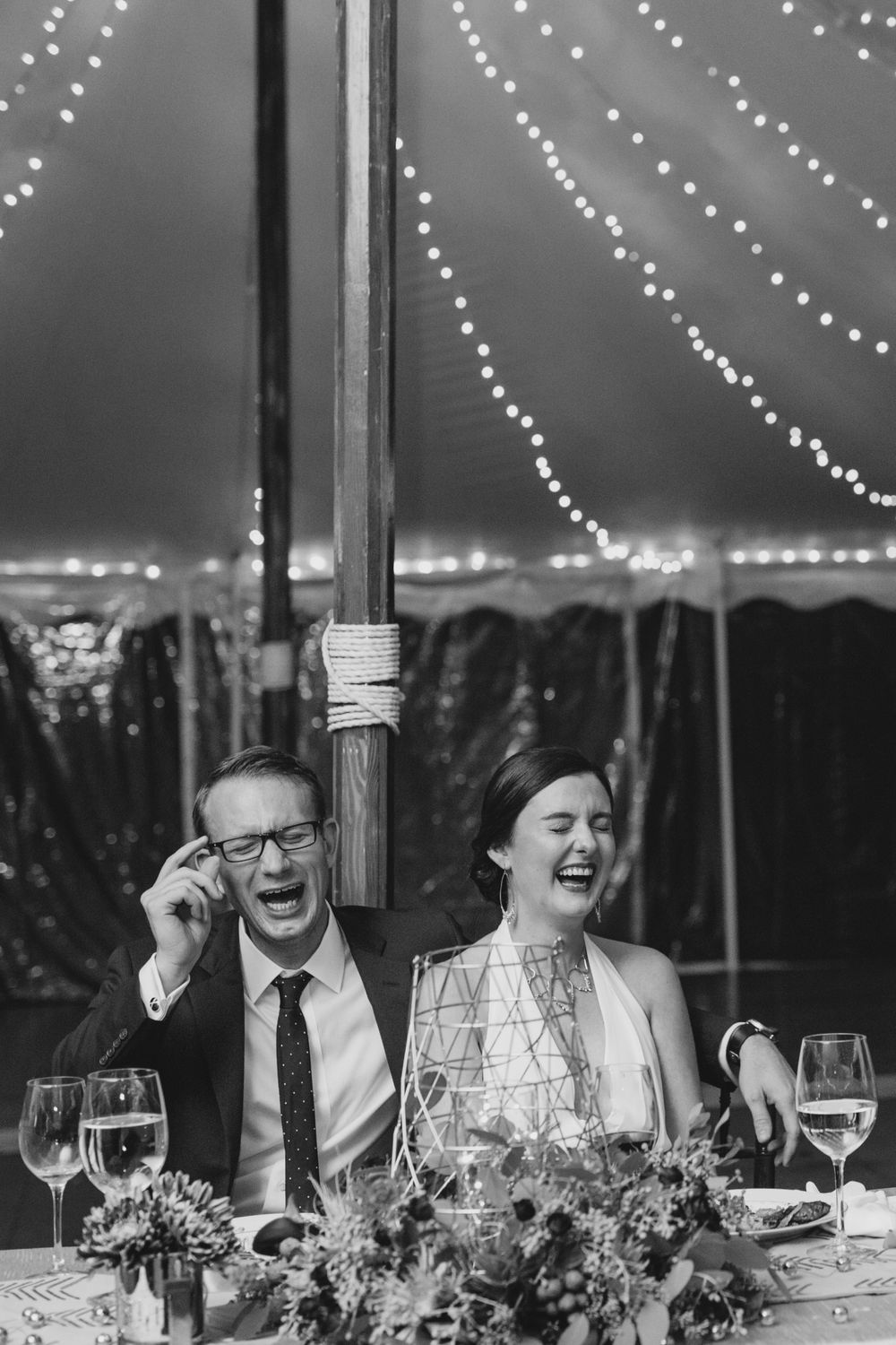 A candid black and white photo of reception guests laughing under twinkling tent lights.