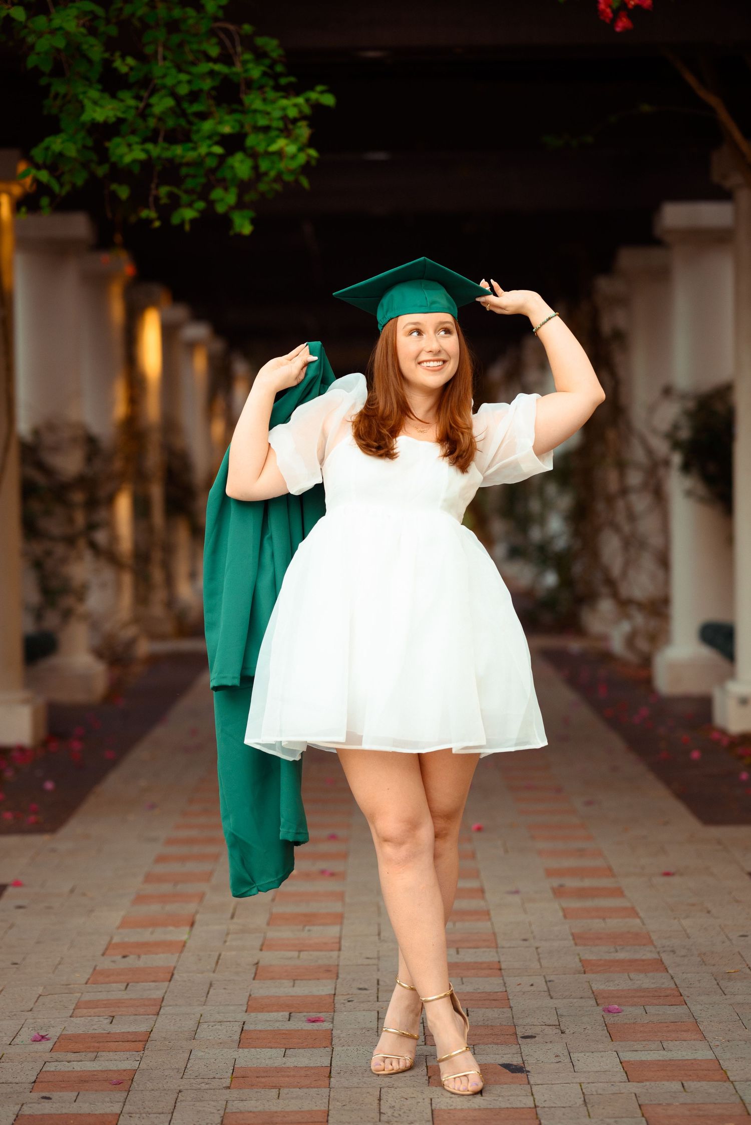 Graduate in white dress and green cap holds graduation gown while posing on brick pathway near columned building.