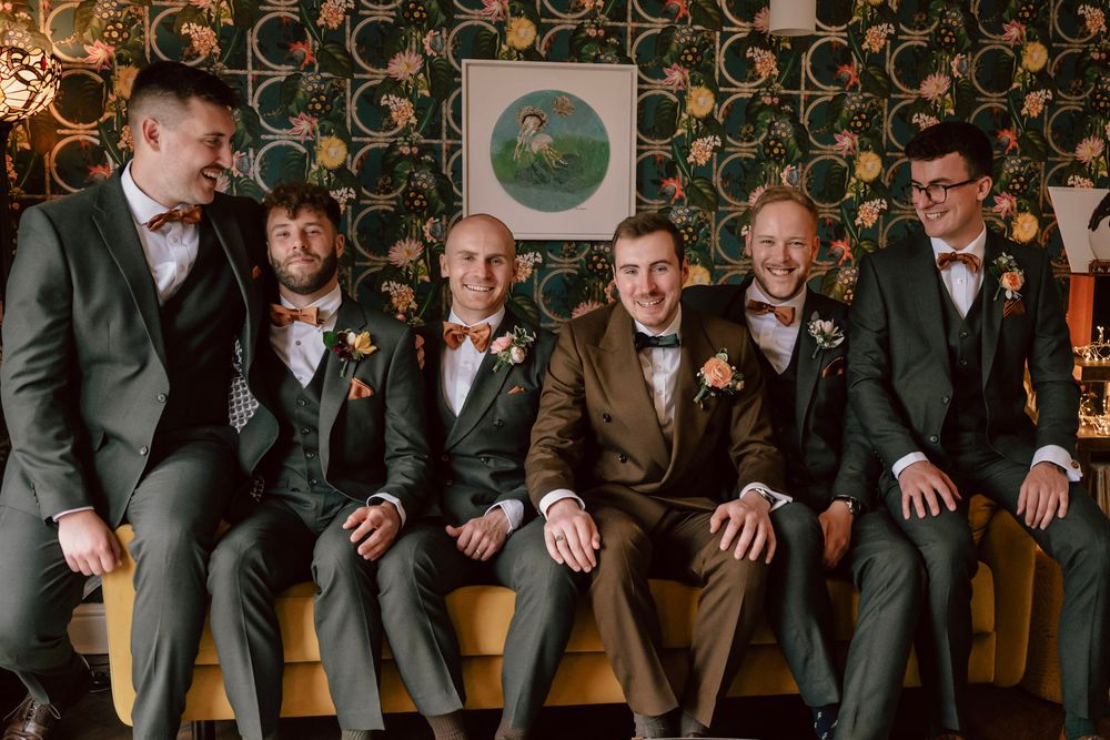 Groomsmen in stylish suits sit together on yellow couch against patterned wallpaper at wedding celebration.